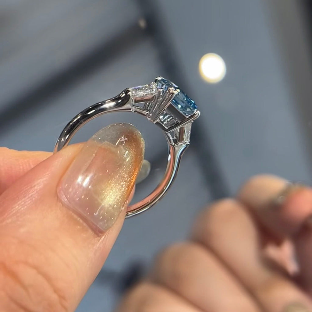 Platinum ring with a blue diamond trilogy ring held by a hand against a neutral background